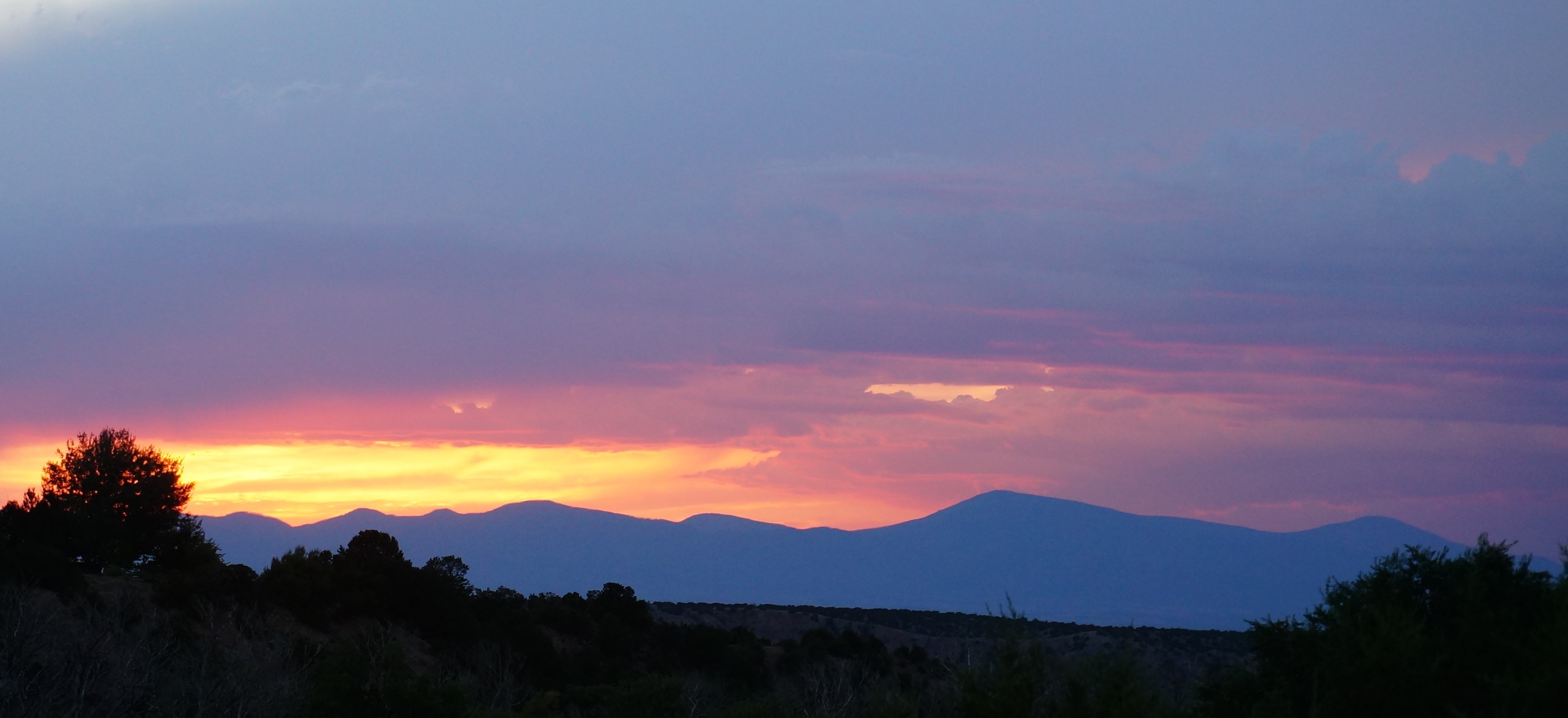 Pink, blue, and gold sunset over the Jemez Mountains with silhouette of a tree-lined ridge